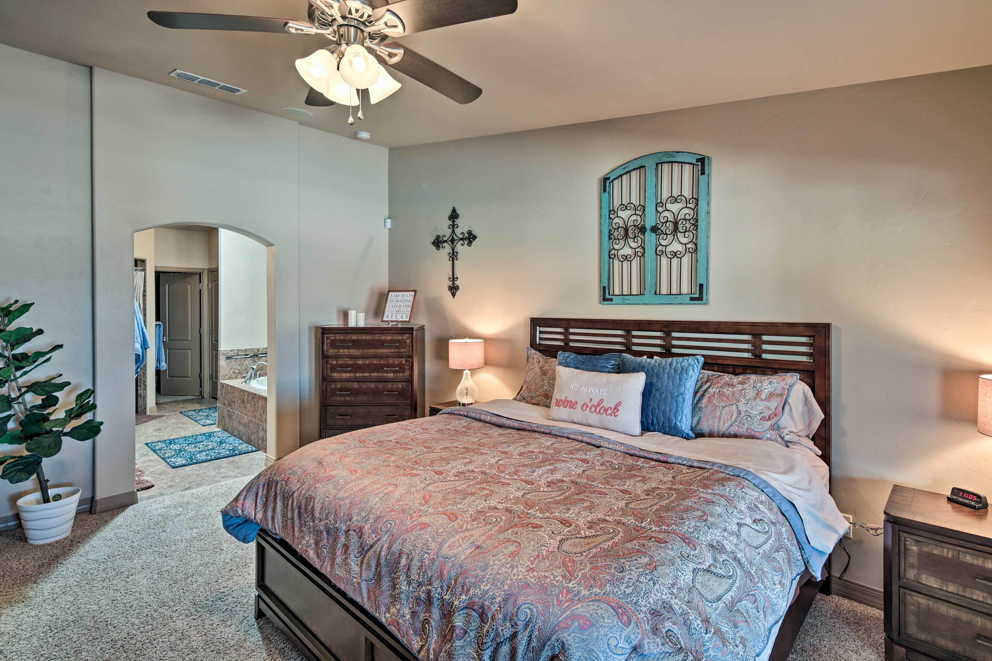 A bedroom with a large bed, a ceiling fan, a nightstand, a dresser, a wall-mounted decorative item, and a door leading to another room.