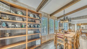 A dining area with a wooden table and chairs, a wooden bookshelf with various items, and a view of the outdoors through large windows. - Ruidoso Home w/ Hot Tub & Sierra Blanca Views (Ruidoso)