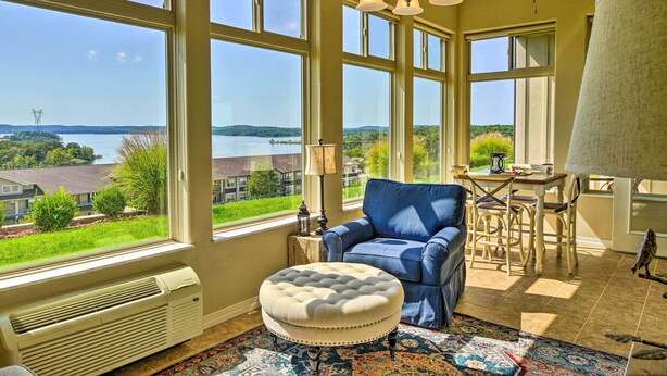 A room with large windows, a blue armchair, a white ottoman, a dining table with chairs, and a ceiling fan. - Branson Condo w/ Table Rock Lake Views, Near Strip (Branson)