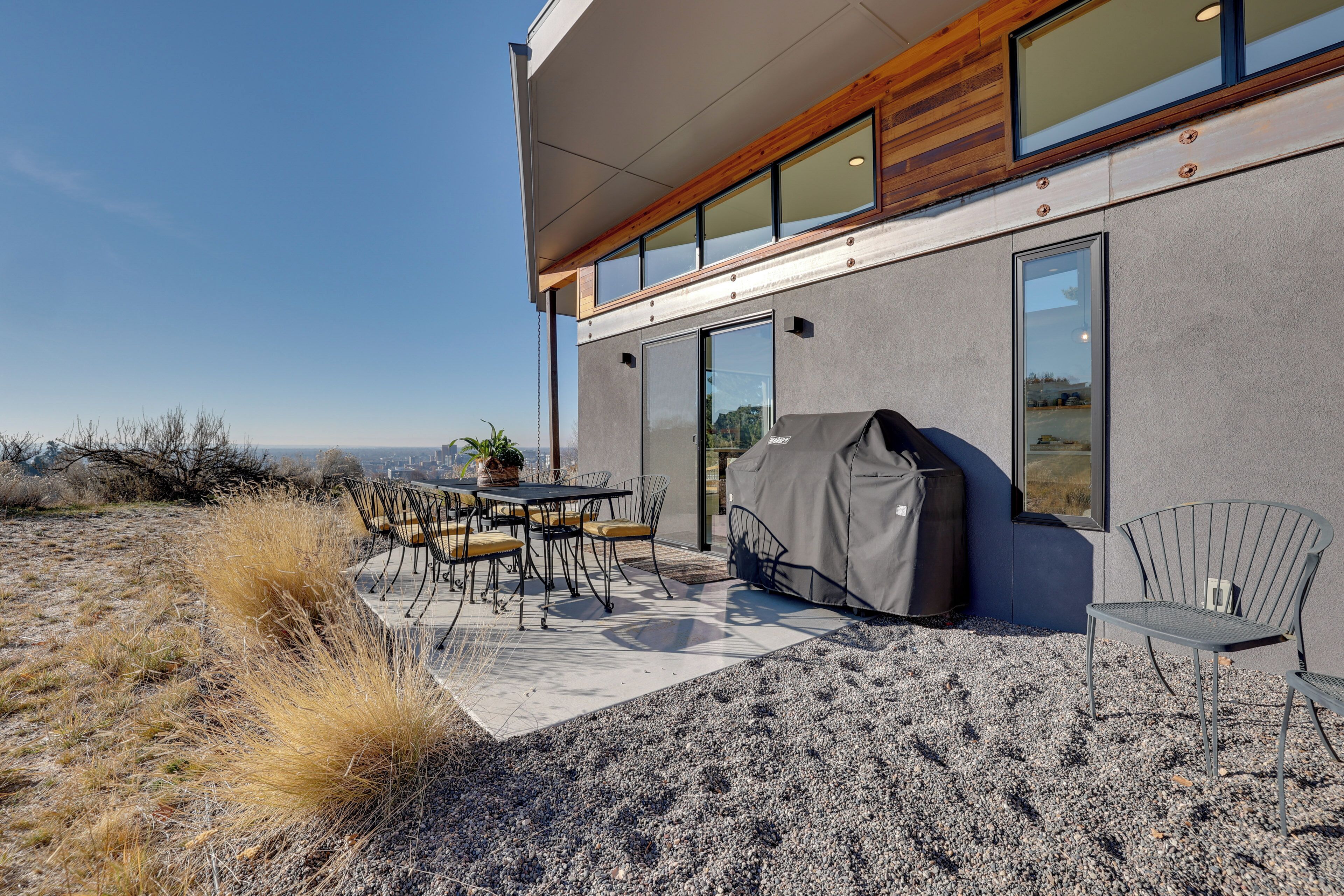 A modern outdoor patio with a table and chairs, a covered grill, and a view of the city.