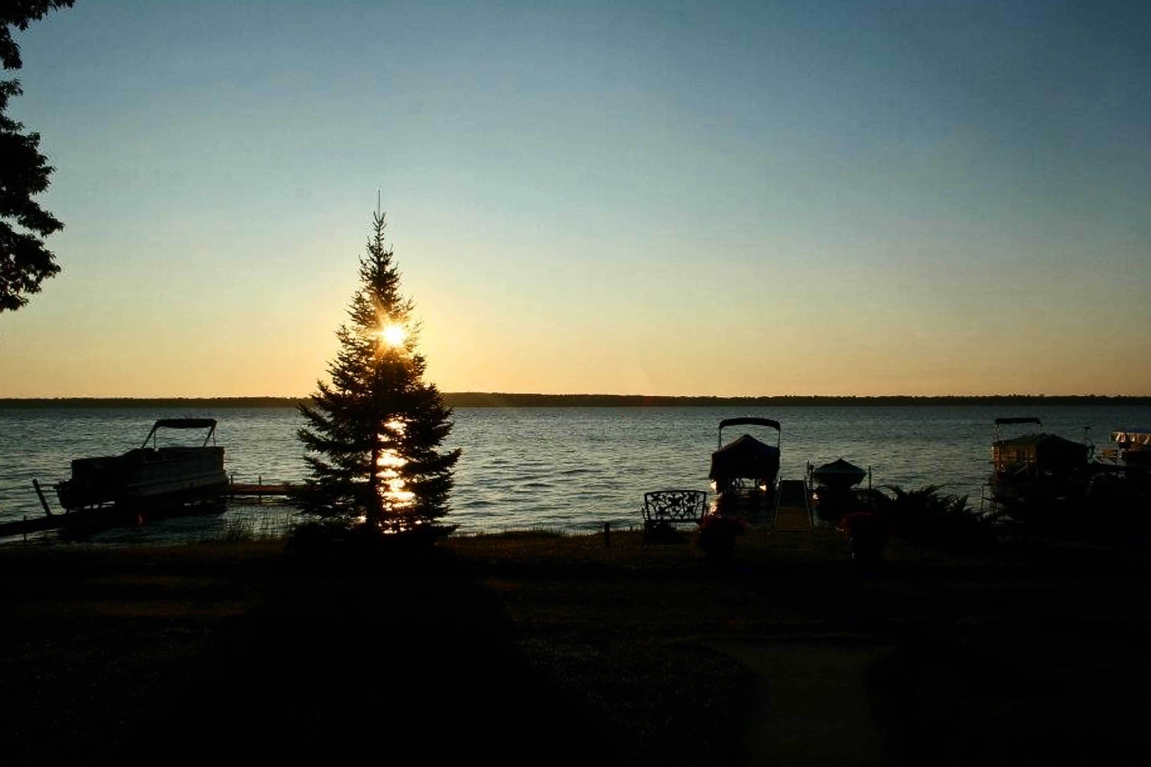 A lakeside view with a boat docked on the left, a tree in the foreground, and a clear sky.