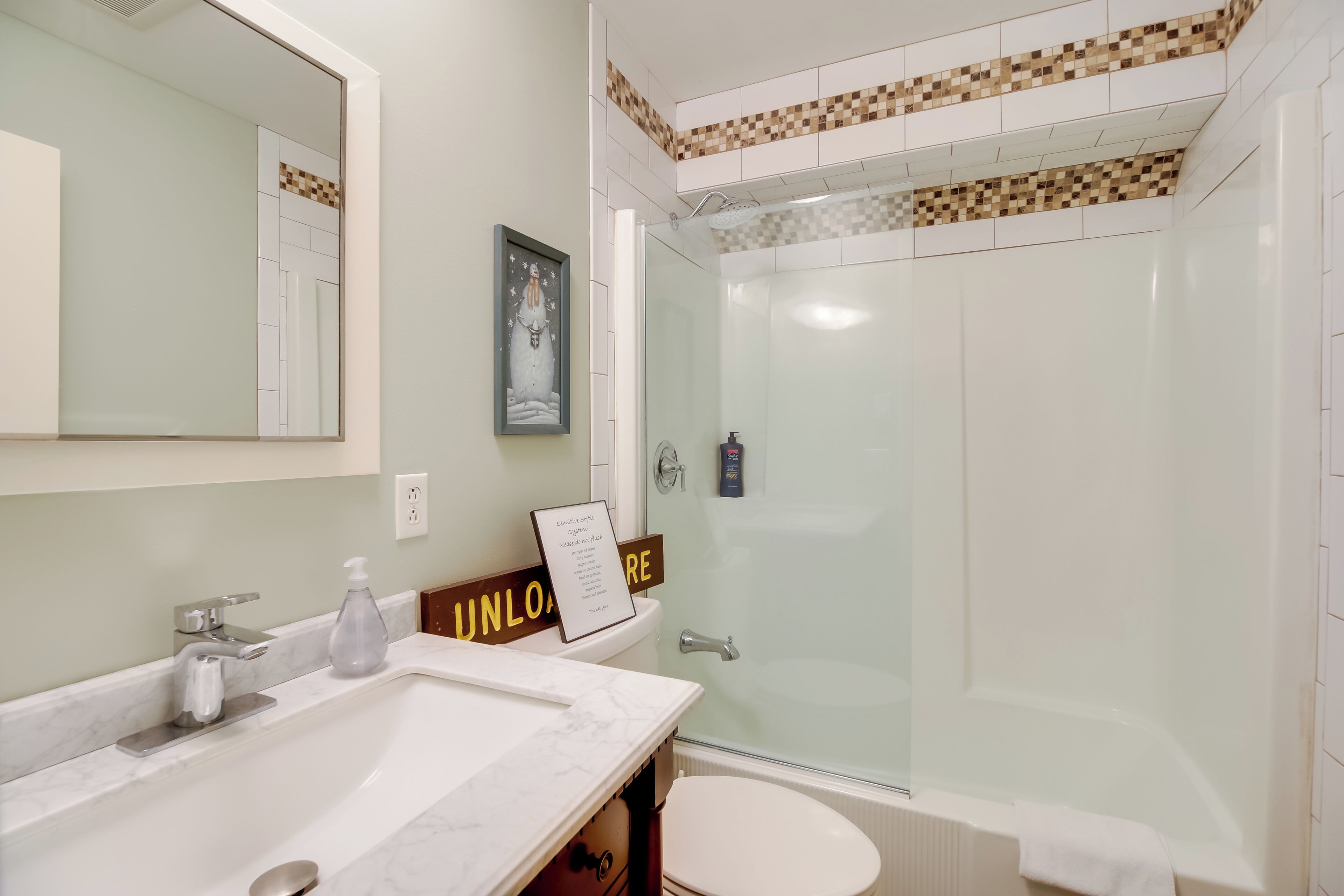 Een badkamer met een wit wastafel, spiegel en toilet. Er is een douche met een glazen deur en een bordje aan de muur.