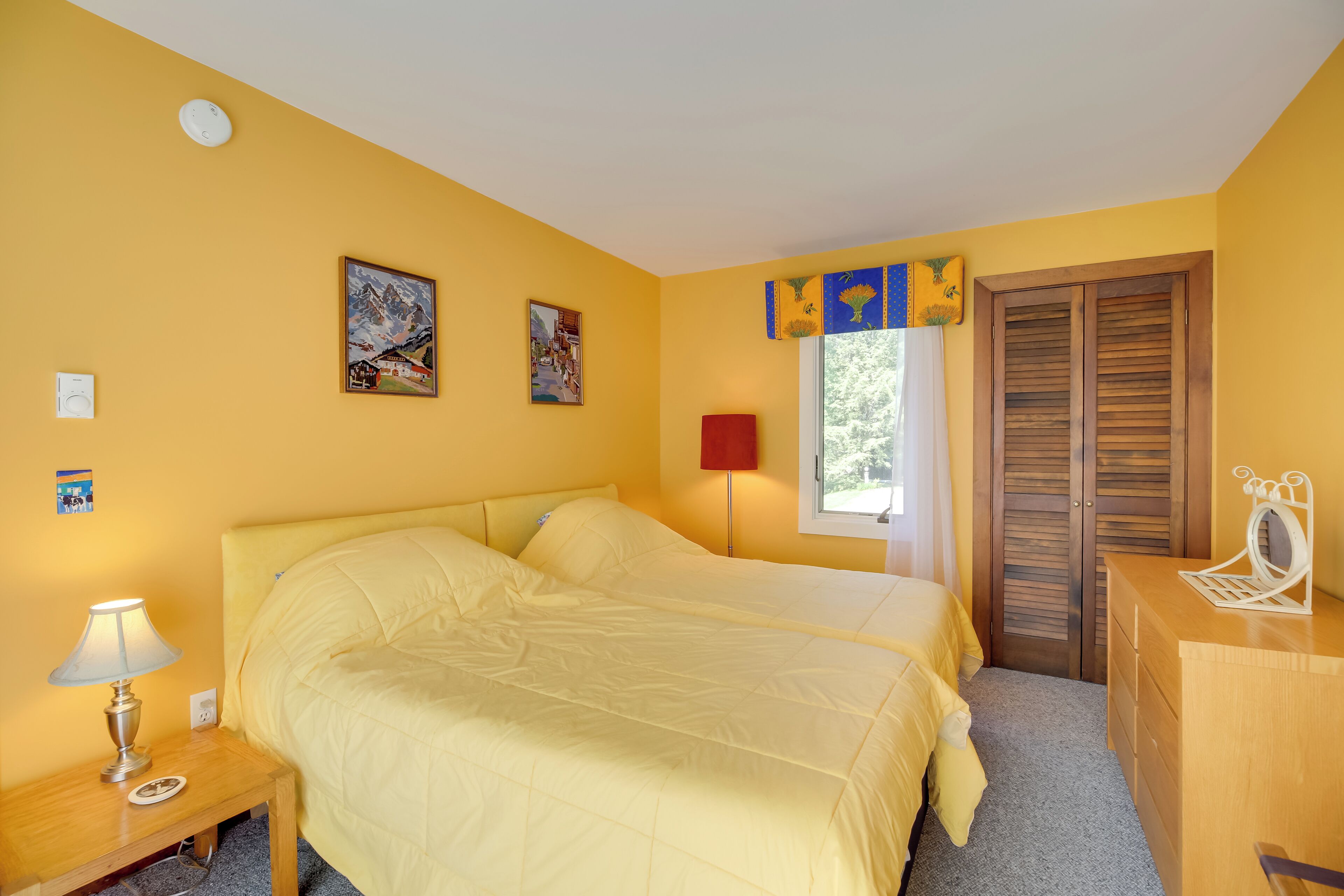 Une chambre avec deux lits, une table de chevet avec une lampe, une commode, une fenêtre avec des volets et deux tableaux accrochés au mur.