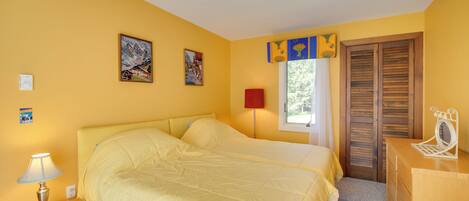 Une chambre avec deux lits, une table de chevet avec une lampe, une commode, une fenêtre avec des volets et deux tableaux accrochés au mur.