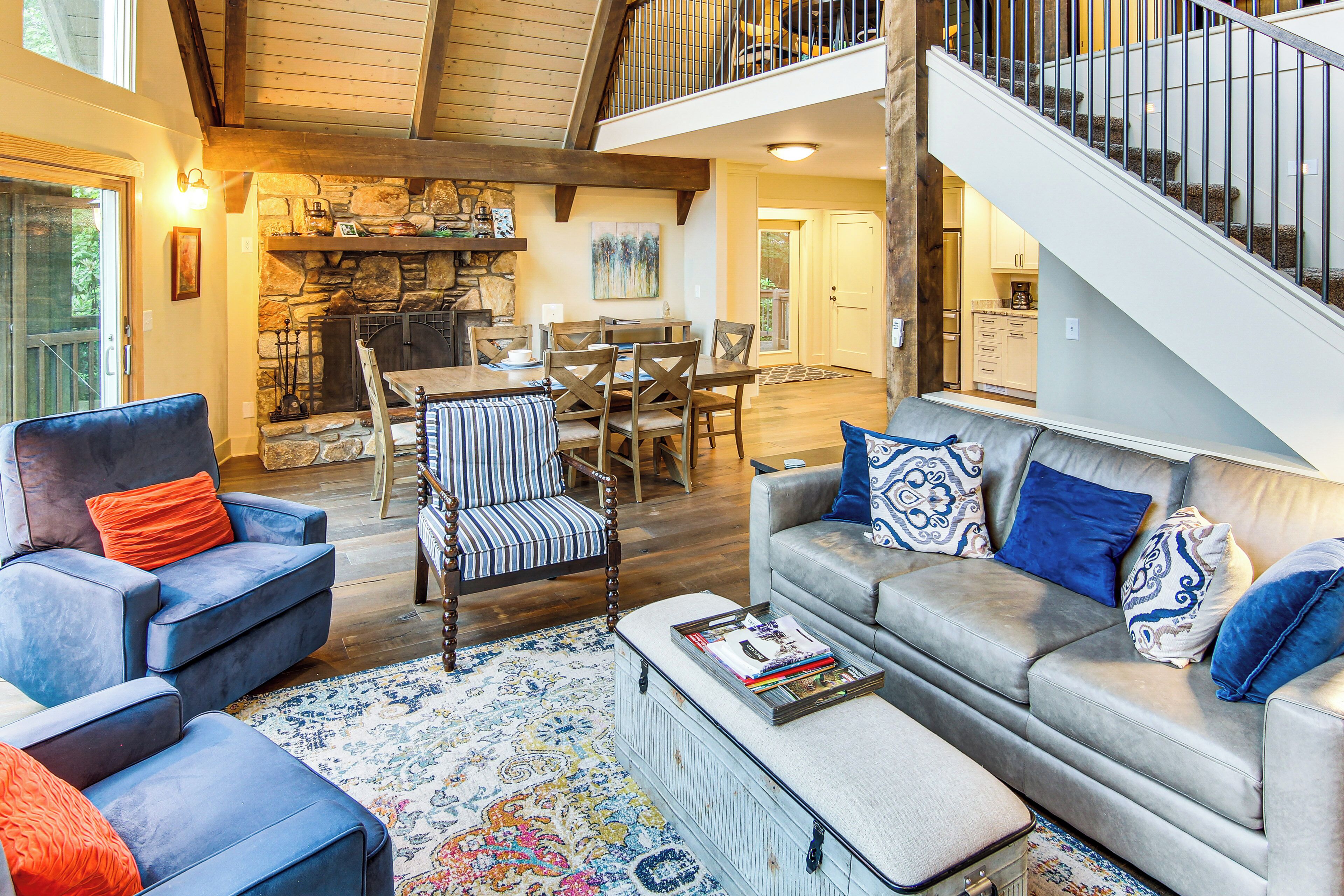 A spacious living area with a stone fireplace, a dining table, and a staircase leading to an upper level.