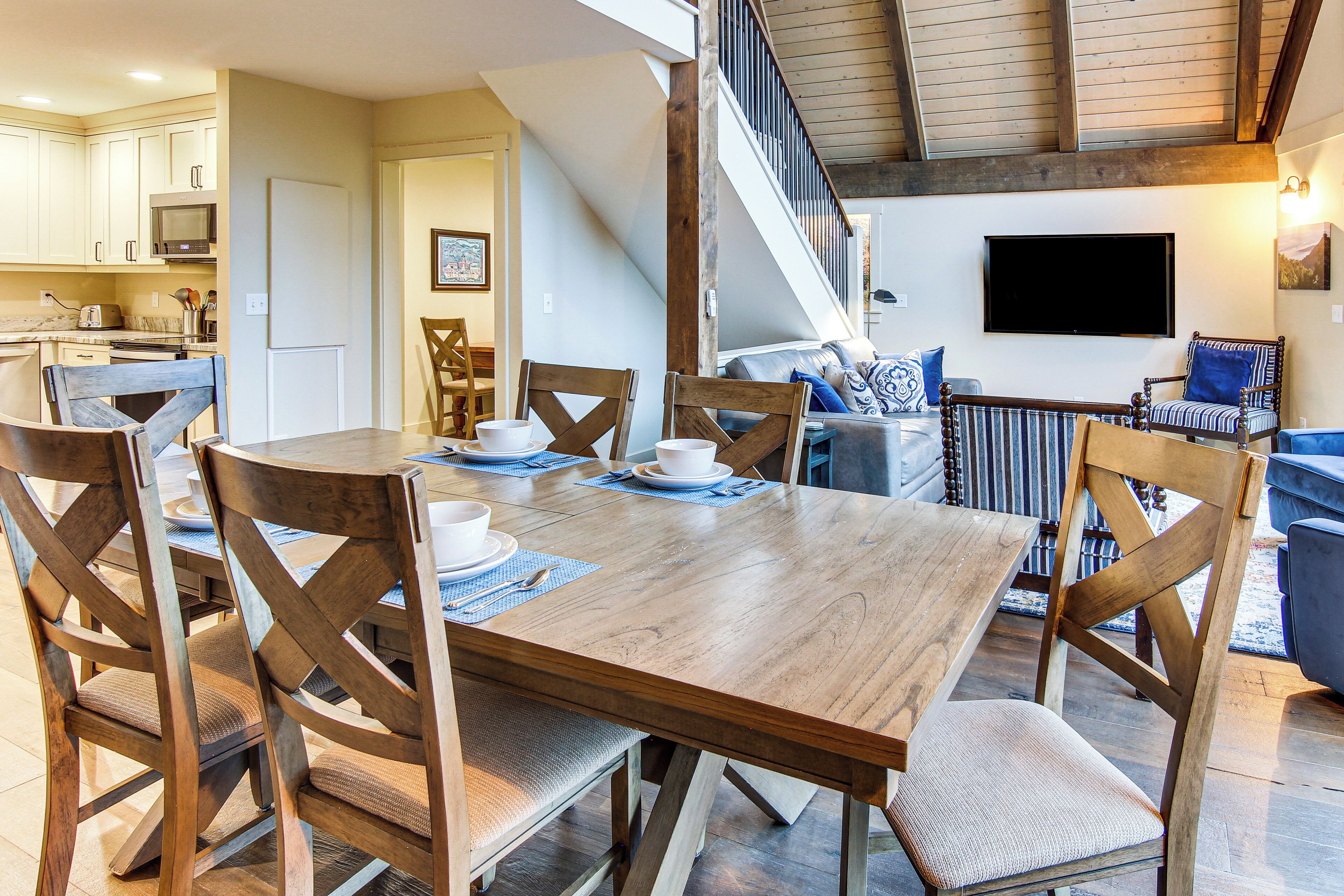 A dining area with a wooden table and chairs, a kitchen in the background, and a staircase leading to another level.