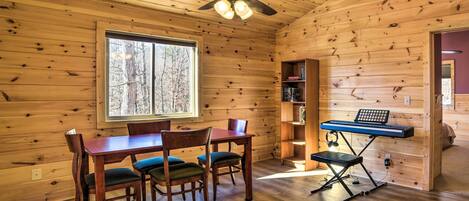 A wooden cabin interior with a dining table, chairs, a ceiling fan, and a keyboard on a stand.