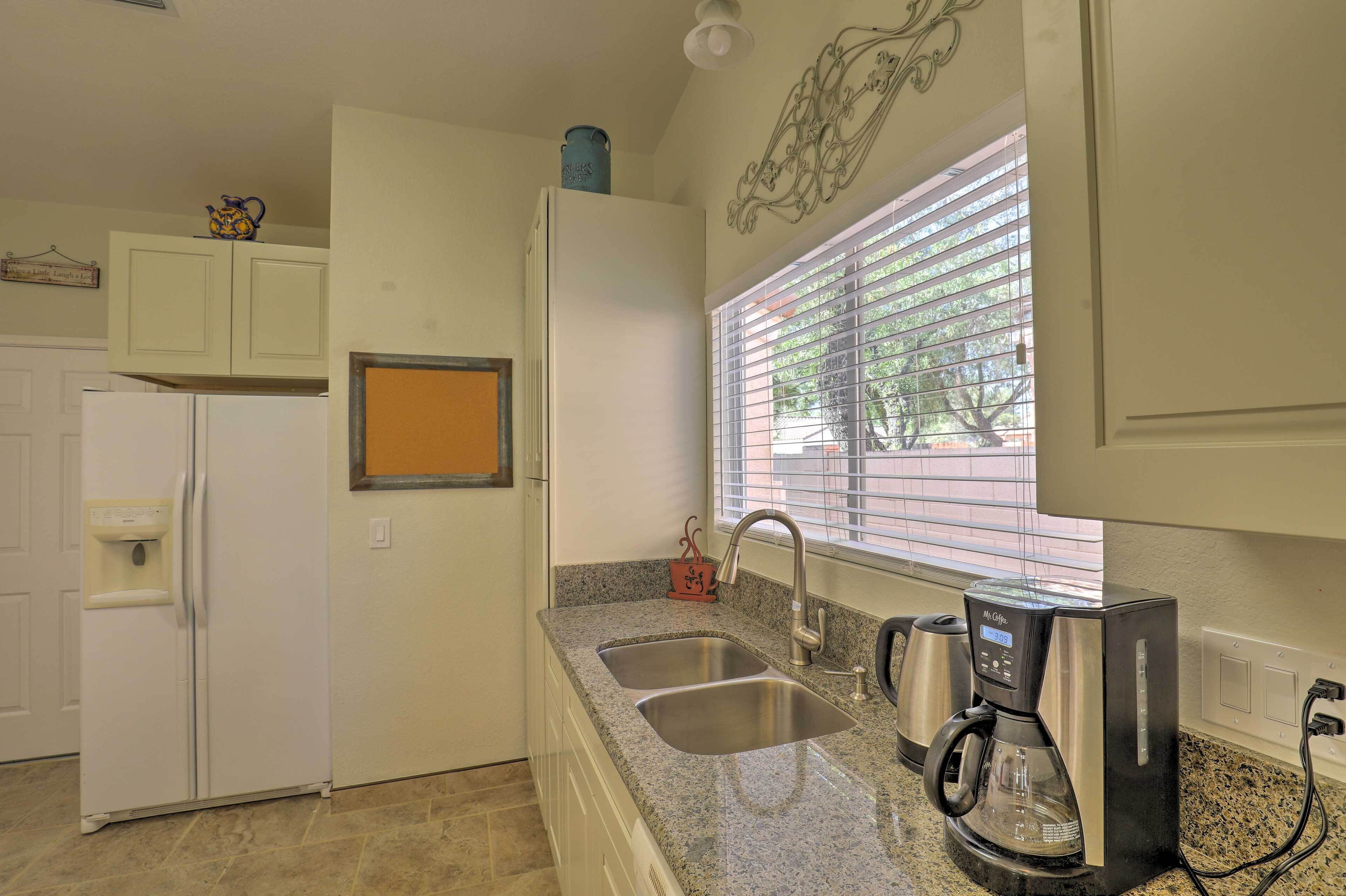 A kitchen with a refrigerator, double sink, coffee maker, and a window with blinds.