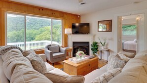 A living room with a fireplace, a television, and a large window with a view of the forest.