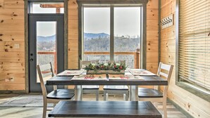 A wooden cabin interior with a dining table set for four, a large window with a mountain view, and a door with a coat rack beside it. - Smoky Mountain Hideaway w/ Hot Tub, Deck & Views! (Gatlinburg)