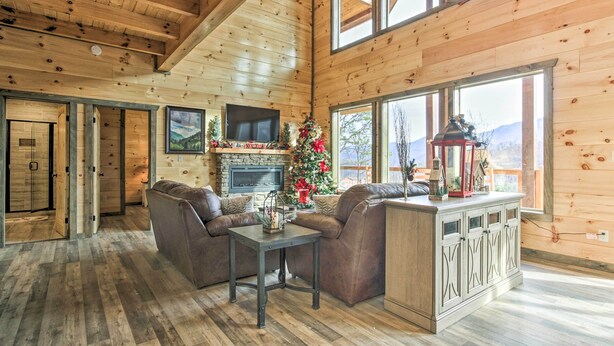 A cozy cabin interior with a fireplace, leather sofas, a dining table, and a large window offering a mountain view. - Smoky Mountain Hideaway w/ Hot Tub, Deck & Views! (Gatlinburg)