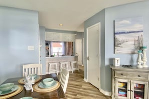 A dining area with a wooden table set for four, a kitchen in the background, and a decorative painting on the wall. - Resort-style Corpus Christi Condo w/ Ocean View! (Corpus Christi)