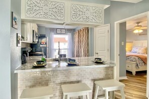 A kitchen with a central island, white bar stools, and a view into a bedroom with a bed and curtains.