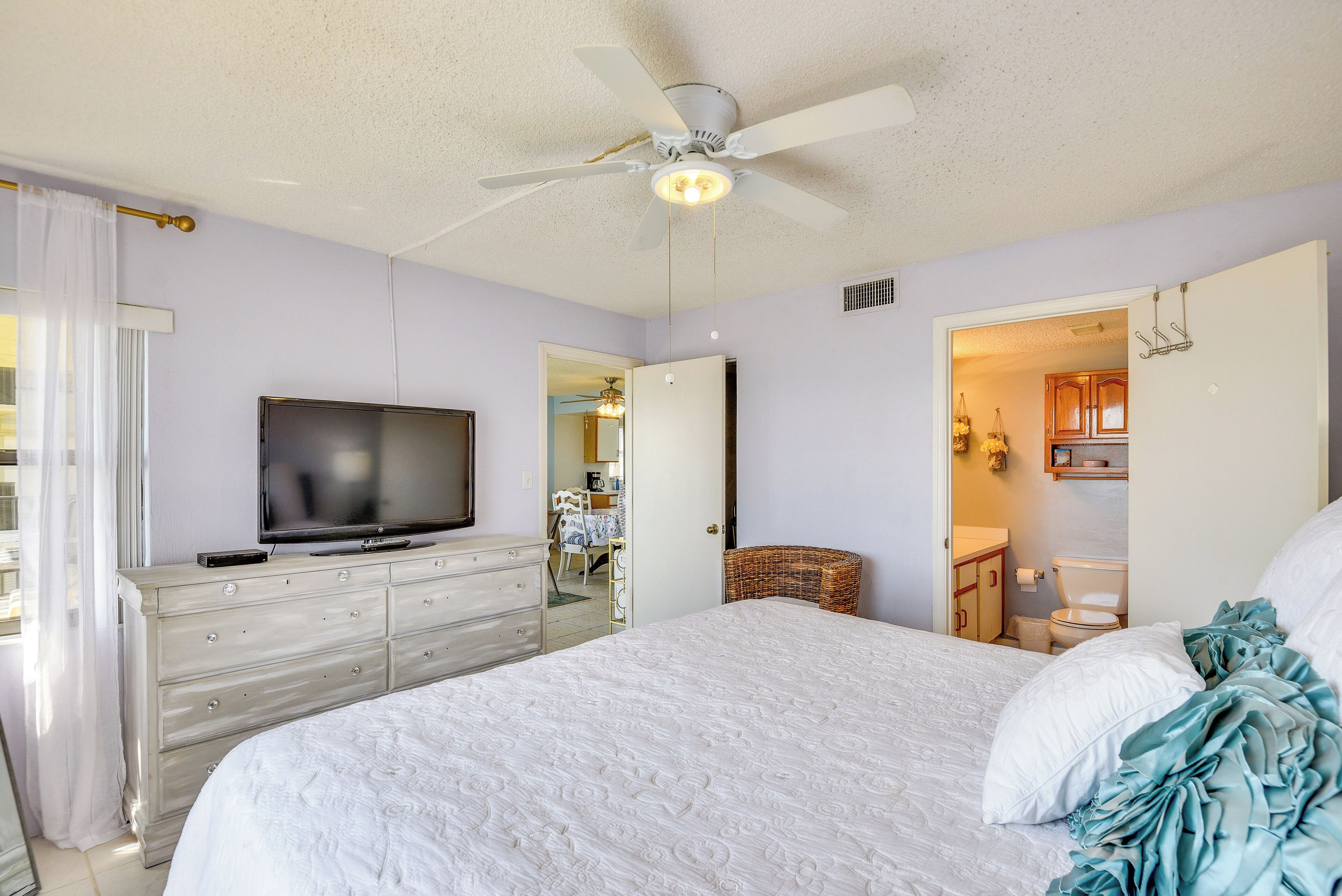 A bedroom with a bed, a dresser, a television, a ceiling fan, and a bathroom visible through an open door.