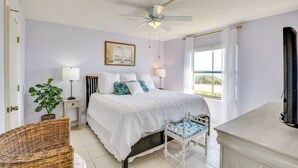 Une chambre à coucher avec un lit, des lampes de chevet, un ventilateur de plafond, une fenêtre avec des rideaux, une plante et un fauteuil en osier.