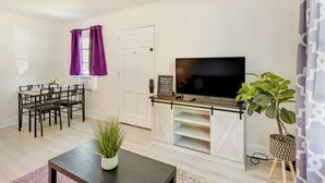 A living room with a TV, dining table, chairs, purple curtains, and a potted plant. - Winston-salem Vacation Rental ~ 3 Mi to Downtown! (Winston-Salem)