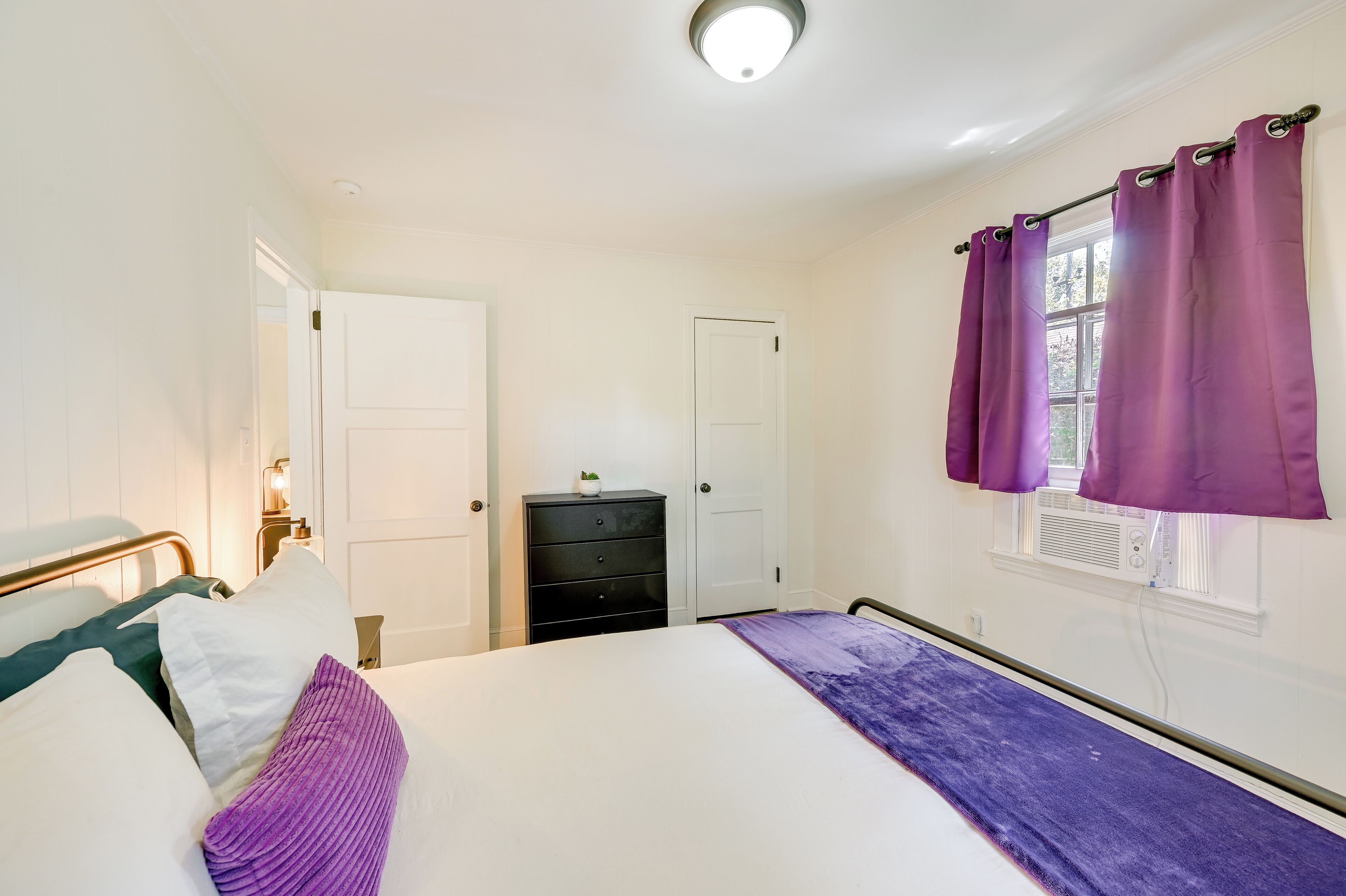 A bedroom with a bed, a dresser, a nightstand, and a window with purple curtains.