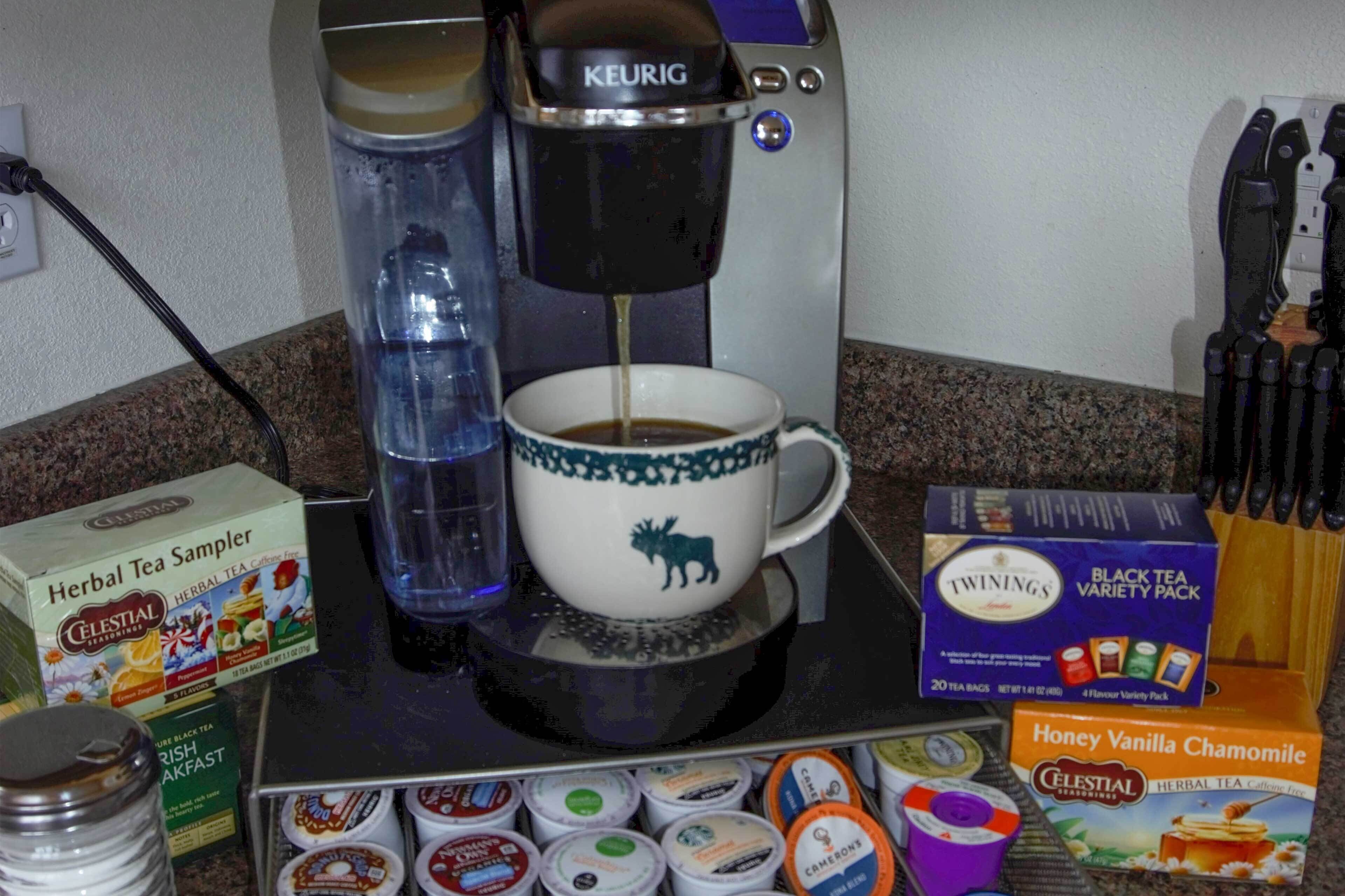 En kaffemaskine med en kop kaffe, en vandflaske og diverse teposer og kaffekapsler på en bordplade.