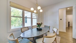 Une salle à manger avec une grande table ovale, six chaises, un lustre et une fenêtre donnant sur un paysage arboré.
