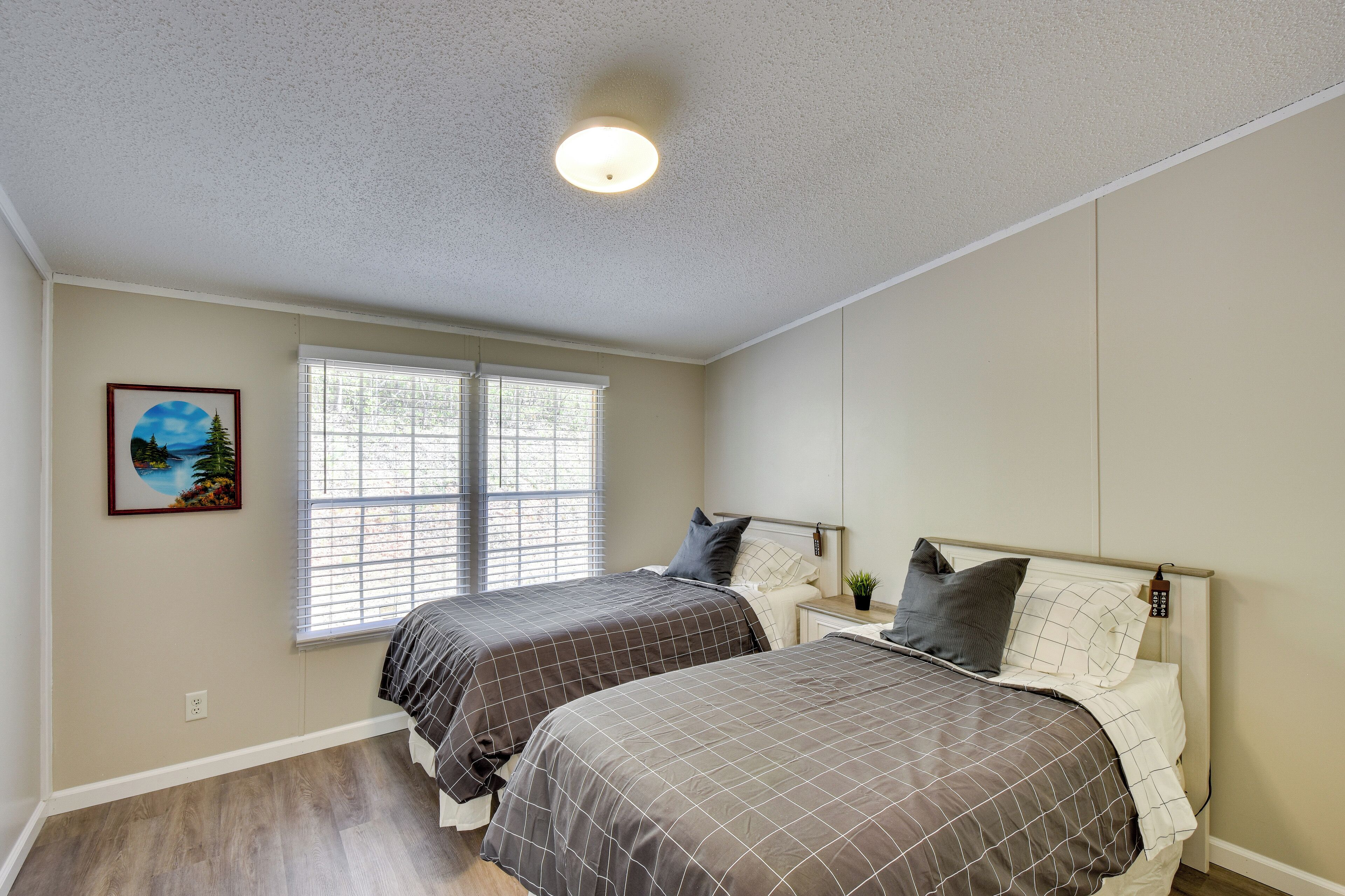 A bedroom with two beds, a window with blinds, a ceiling light, and a framed picture on the wall.