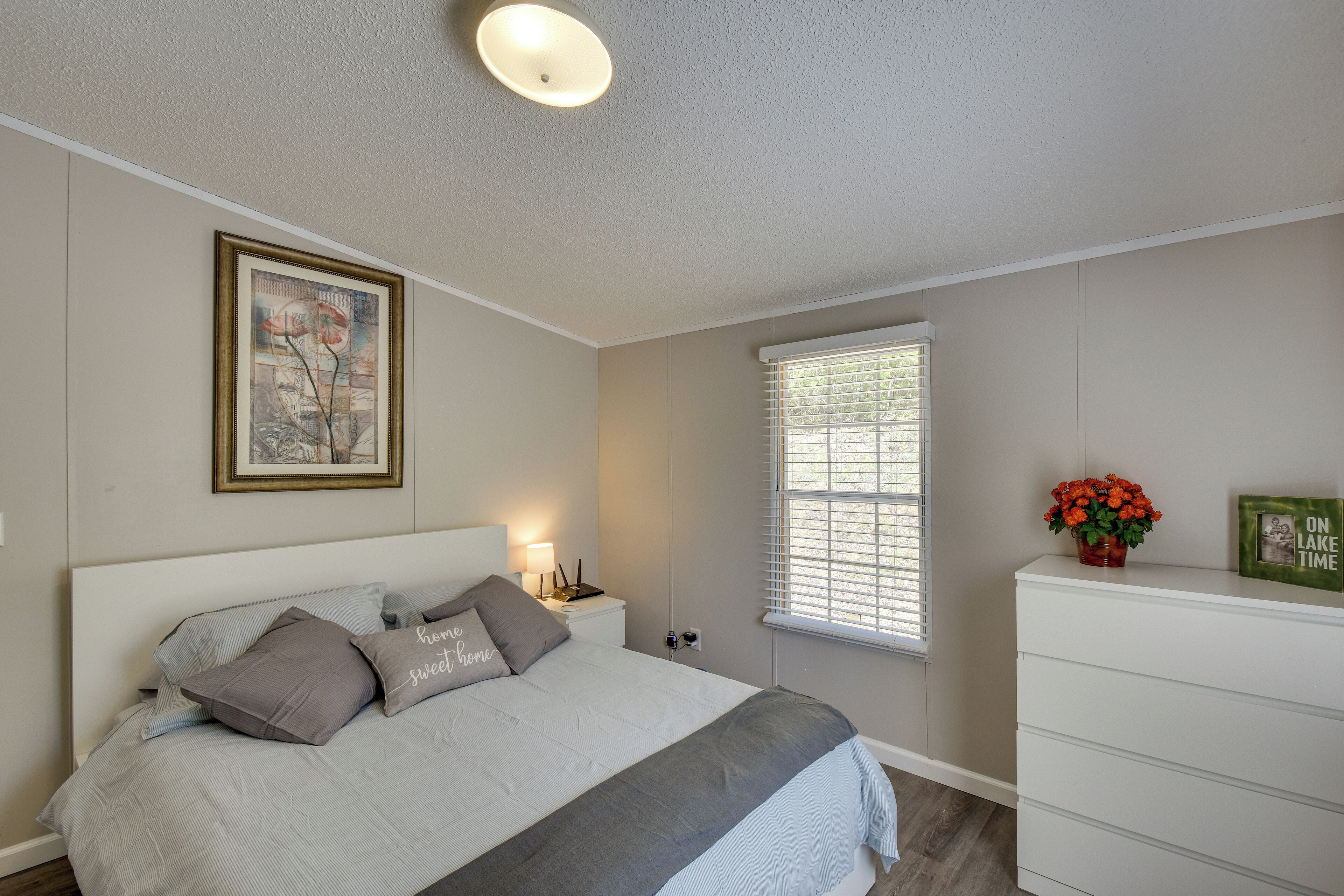 A bedroom with a bed, bedside table, lamp, window, dresser, and a framed picture on the wall.