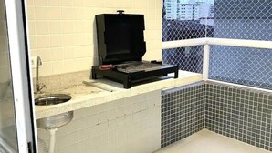Une petite cuisine équipée d’un barbecue portable, d’un évier et d’une fenêtre donnant sur des immeubles.