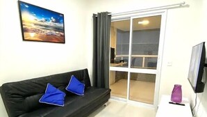 Un salon moderne avec un canapé noir, un meuble TV blanc et une porte vitrée donnant sur un balcon.