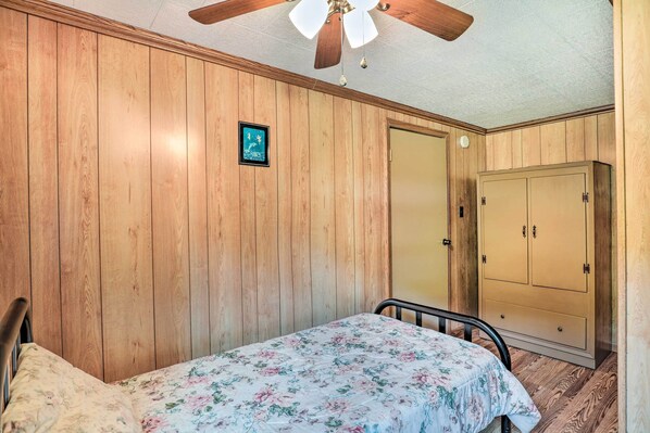 A bedroom with wooden paneling, a bed with a floral bedspread, a ceiling fan, and a wardrobe. - Bonfires & Bbqs! Creekside Retreat in Marshall (Marshall)