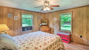 A bedroom with wooden walls, a bed with floral bedding, a ceiling fan, a dresser, a mirror, and a suitcase. - Bonfires & Bbqs! Creekside Retreat in Marshall (Marshall)