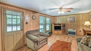 A cozy living room with wooden walls, a ceiling fan, a television, and a fireplace. - Bonfires & Bbqs! Creekside Retreat in Marshall (Marshall)