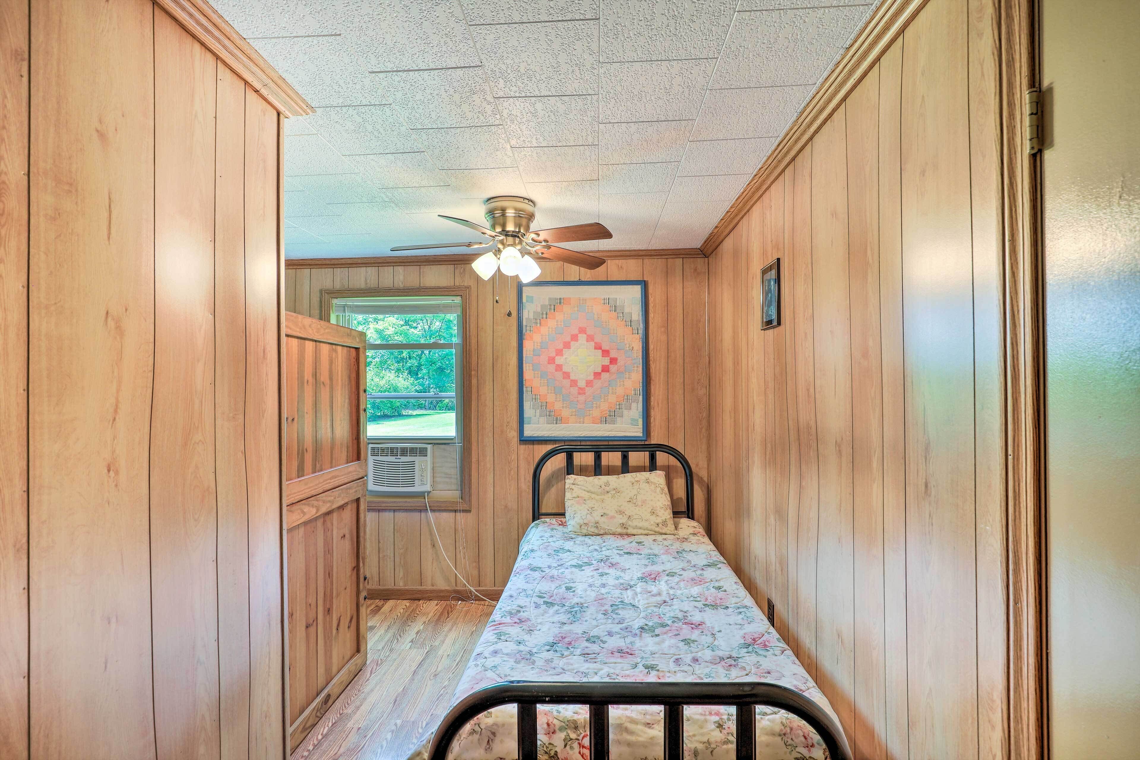 A small, single-bed room with wooden walls, a ceiling fan, and a window with a view of greenery.