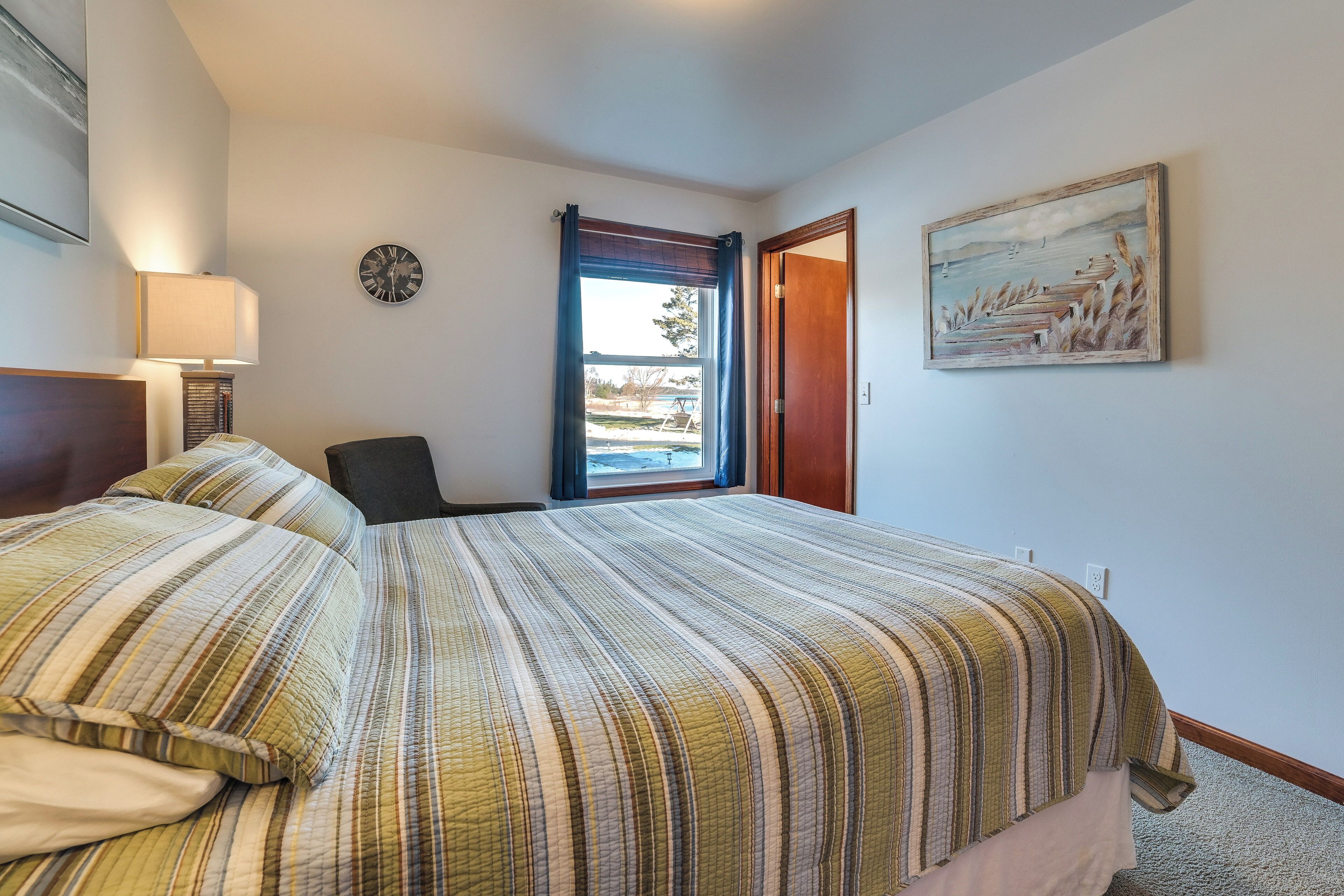 A hotel room with a large bed, a chair, a desk, a lamp, a clock, a painting, and a window with curtains.