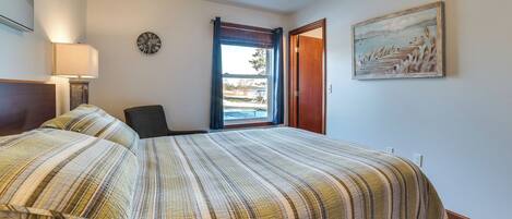 A hotel room with a large bed, a chair, a desk, a lamp, a clock, a painting, and a window with curtains.