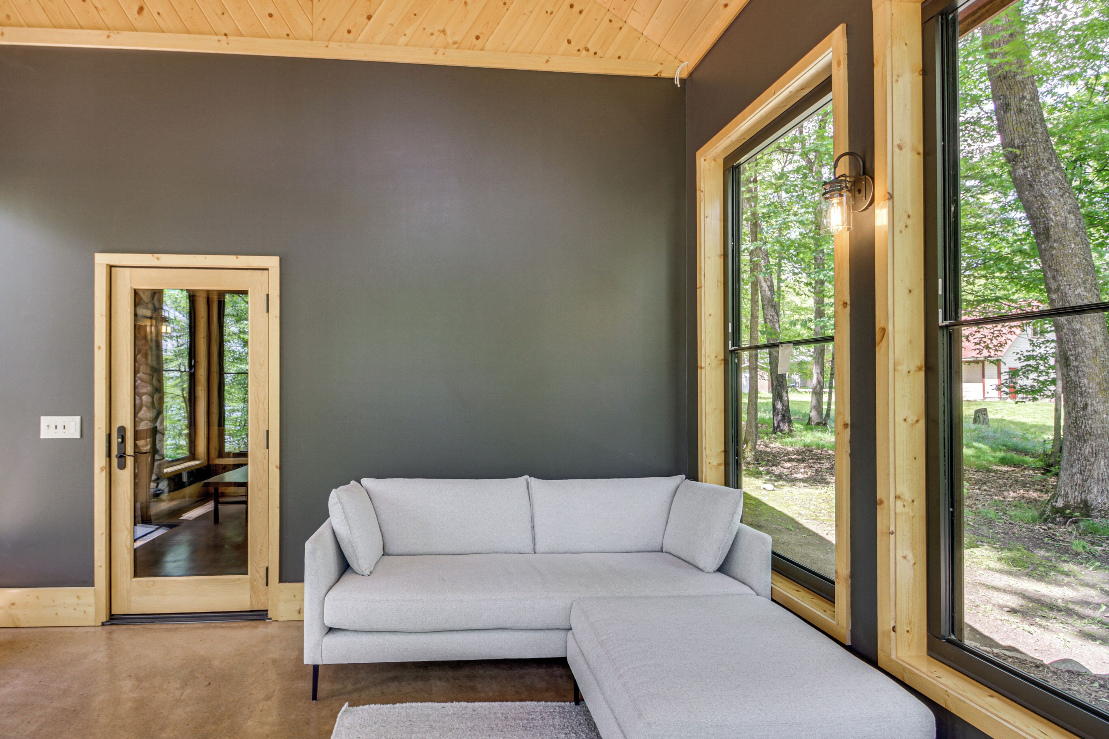 A modern interior with a grey wall, a wooden ceiling, a grey sectional sofa, and a large window with a view of trees.
