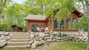 A wooden cabin with a stone staircase, a patio with chairs, and a large window overlooking the forest. - Waterfront Cabin on Trout Lake w/ Private Dock! (Bovey)
