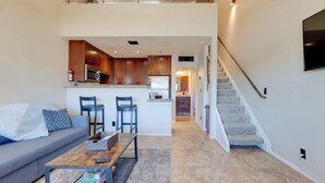 A modern living room with a grey sofa, a coffee table, and two blue chairs. A kitchen area is visible in the background with wooden cabinets and a microwave. - Scottsdale Loft Near Golf Course & Old Town! (Scottsdale)