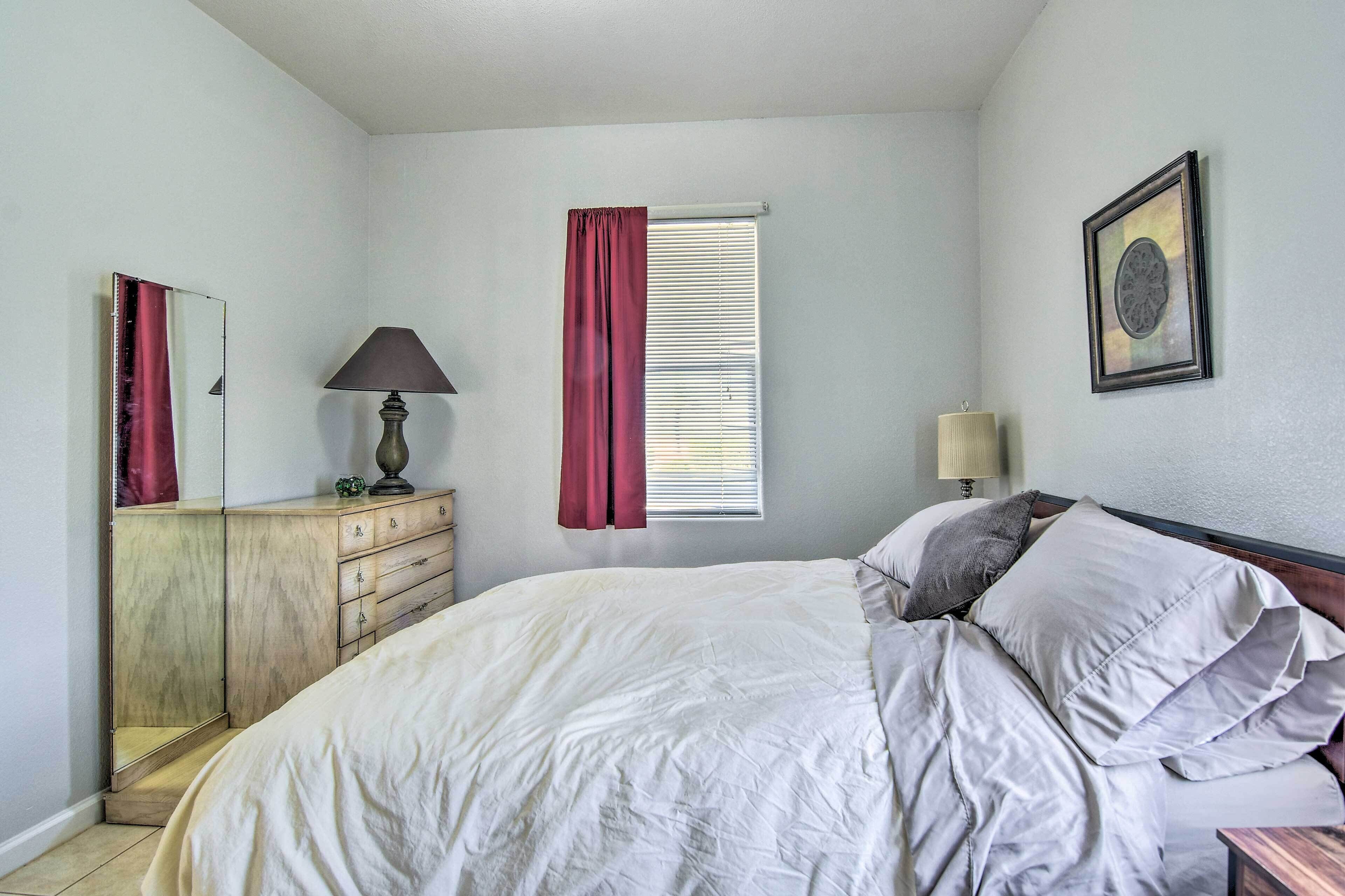 A bedroom with a bed, a dresser, a window with curtains, a lamp, and a framed picture on the wall.