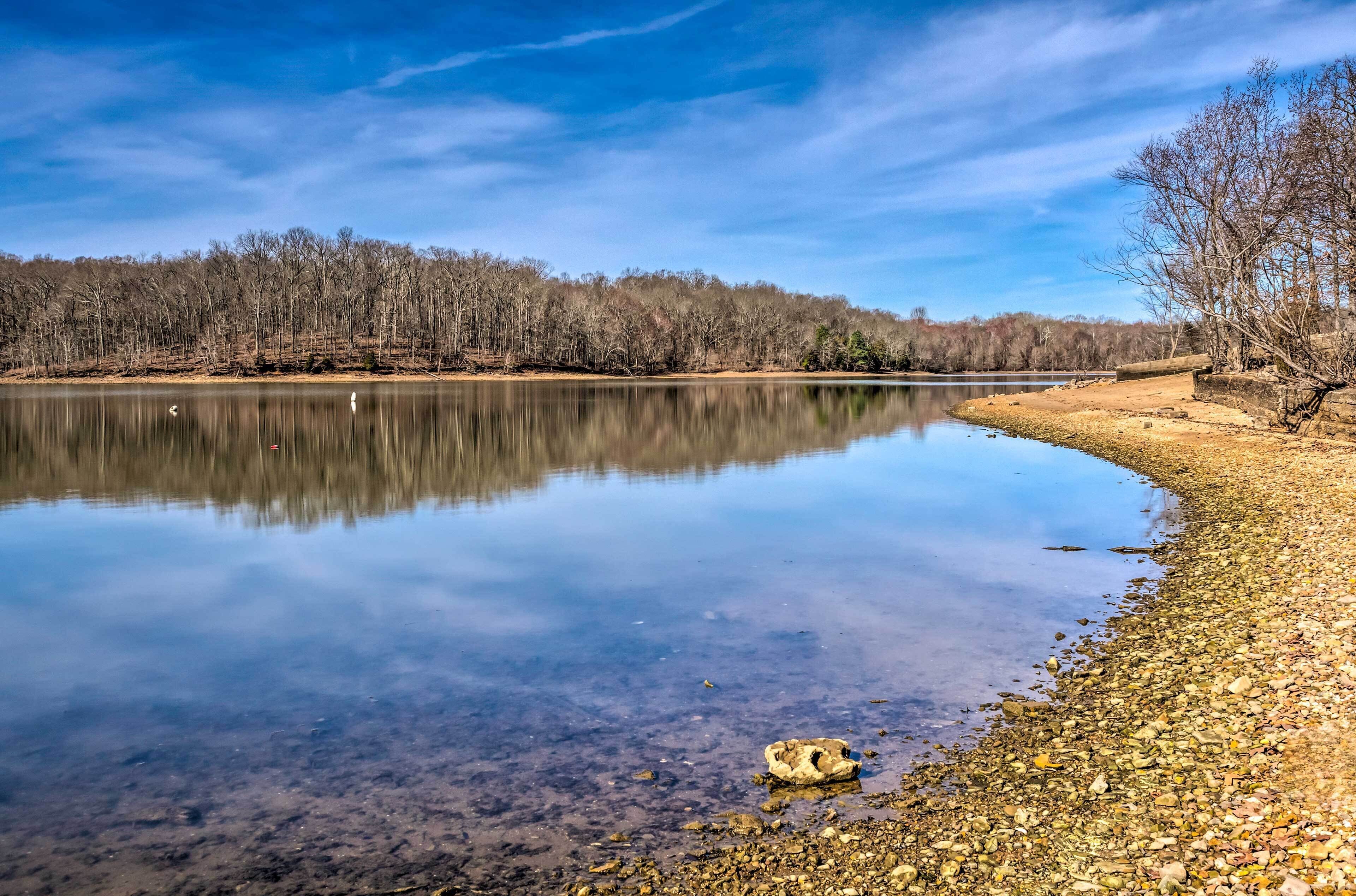 A calm lake with clear reflections, surrounded by a forested area and a rocky shoreline.