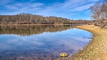 A calm lake with clear reflections, surrounded by a forested area and a rocky shoreline.