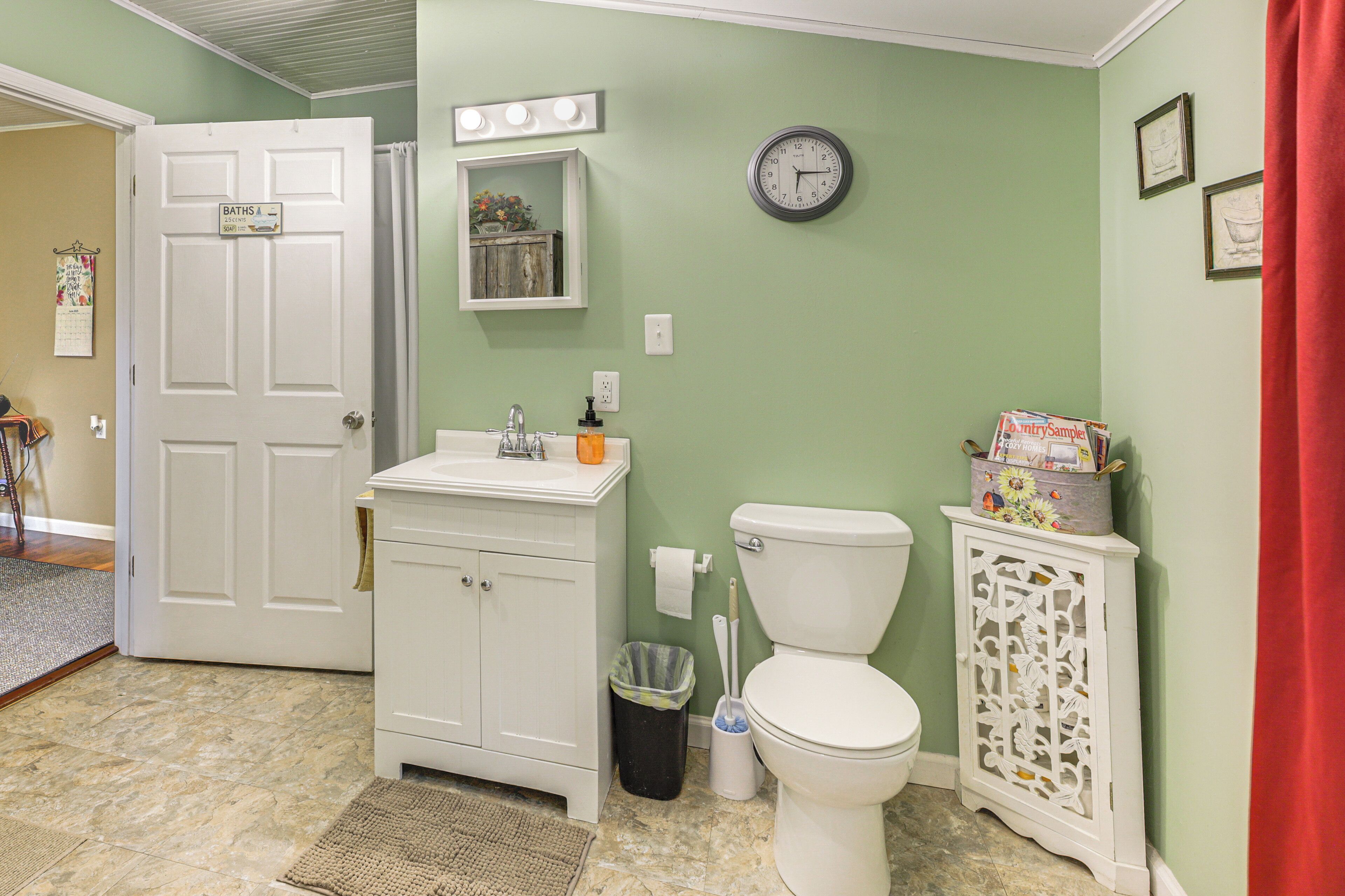 A bathroom with a white door, a sink, a toilet, a clock, and a trash can.