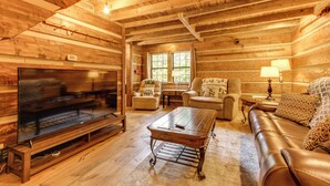 L’intérieur d’une cabane en rondins, avec un plafond en bois, un grand téléviseur à écran plat, un canapé en cuir, une table basse et une fenêtre donnant sur un paysage verdoyant.