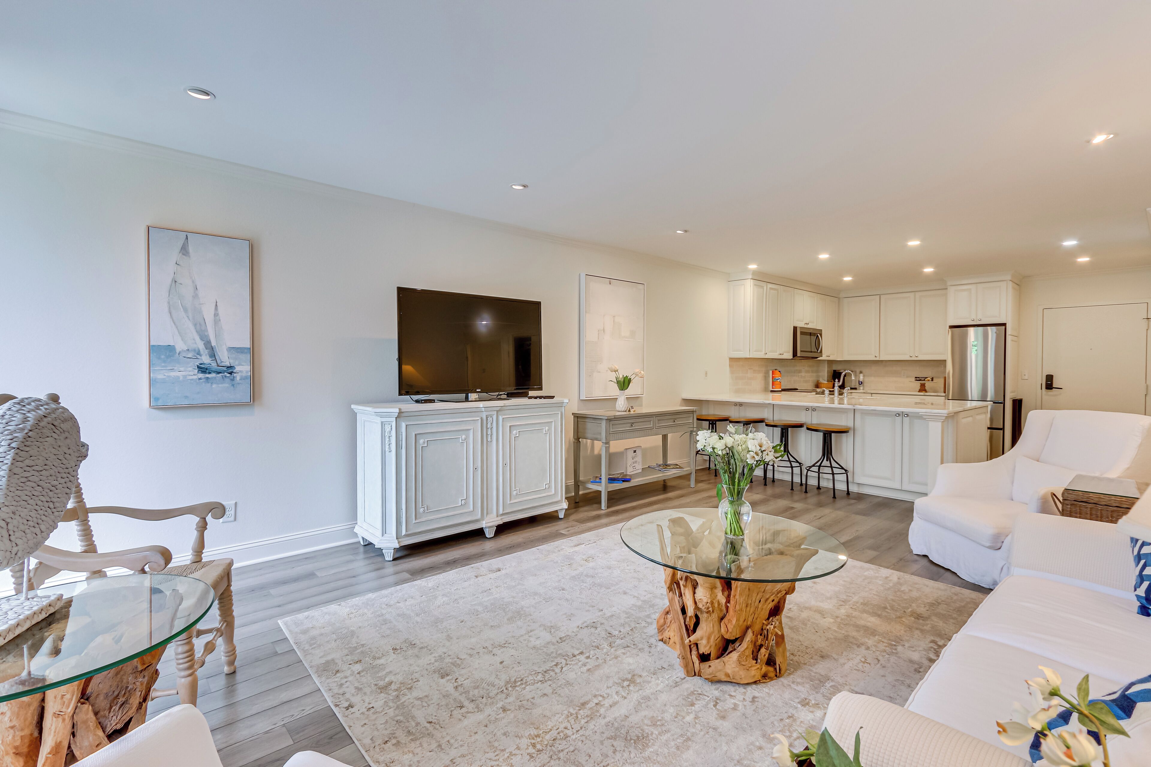 A modern living room with a glass coffee table, white sofas, and a kitchen in the background.