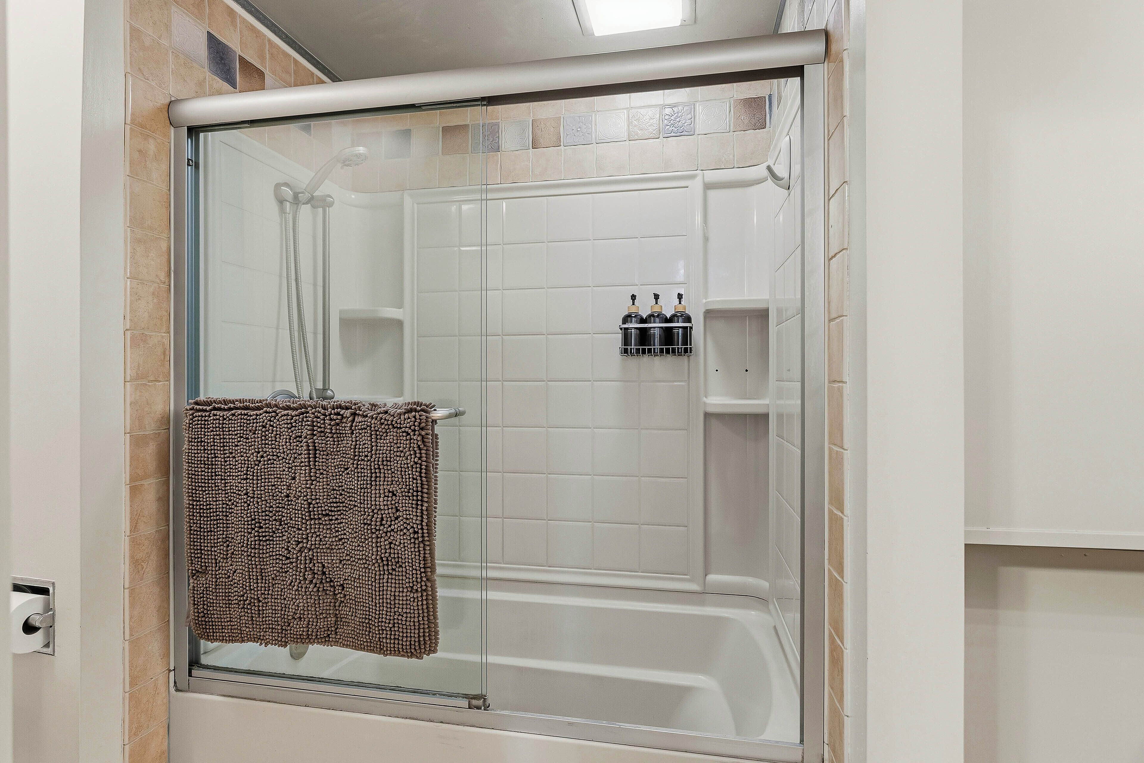 Une douche fermée par des parois de verre, avec un mur recouvert de carreaux blancs, un porte-serviettes avec une serviette et une étagère avec des bouteilles.