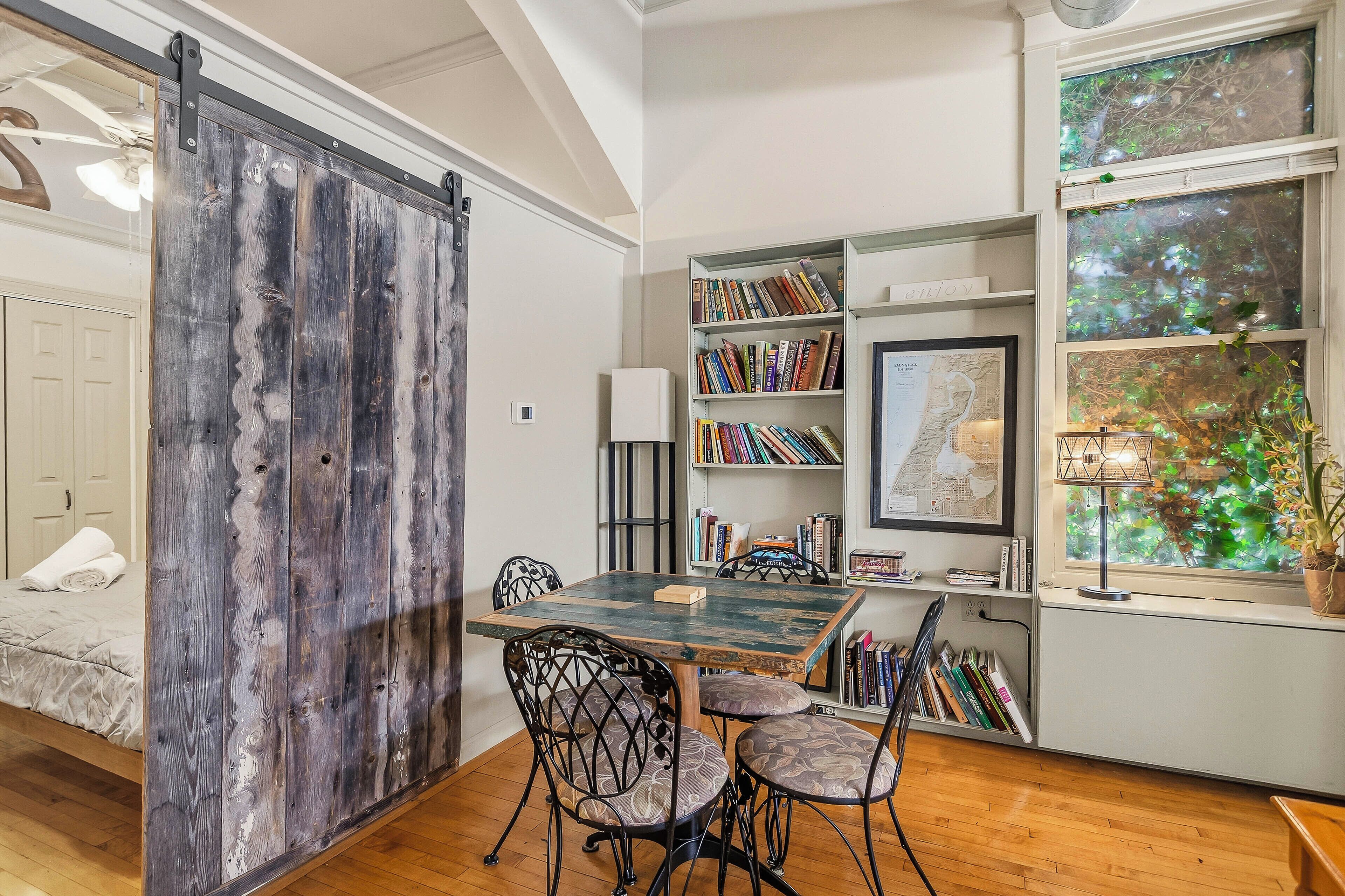 Una habitación con puerta de madera, una mesa de comedor con sillas, una estantería, un mapa en la pared y una ventana con vistas a la vegetación.