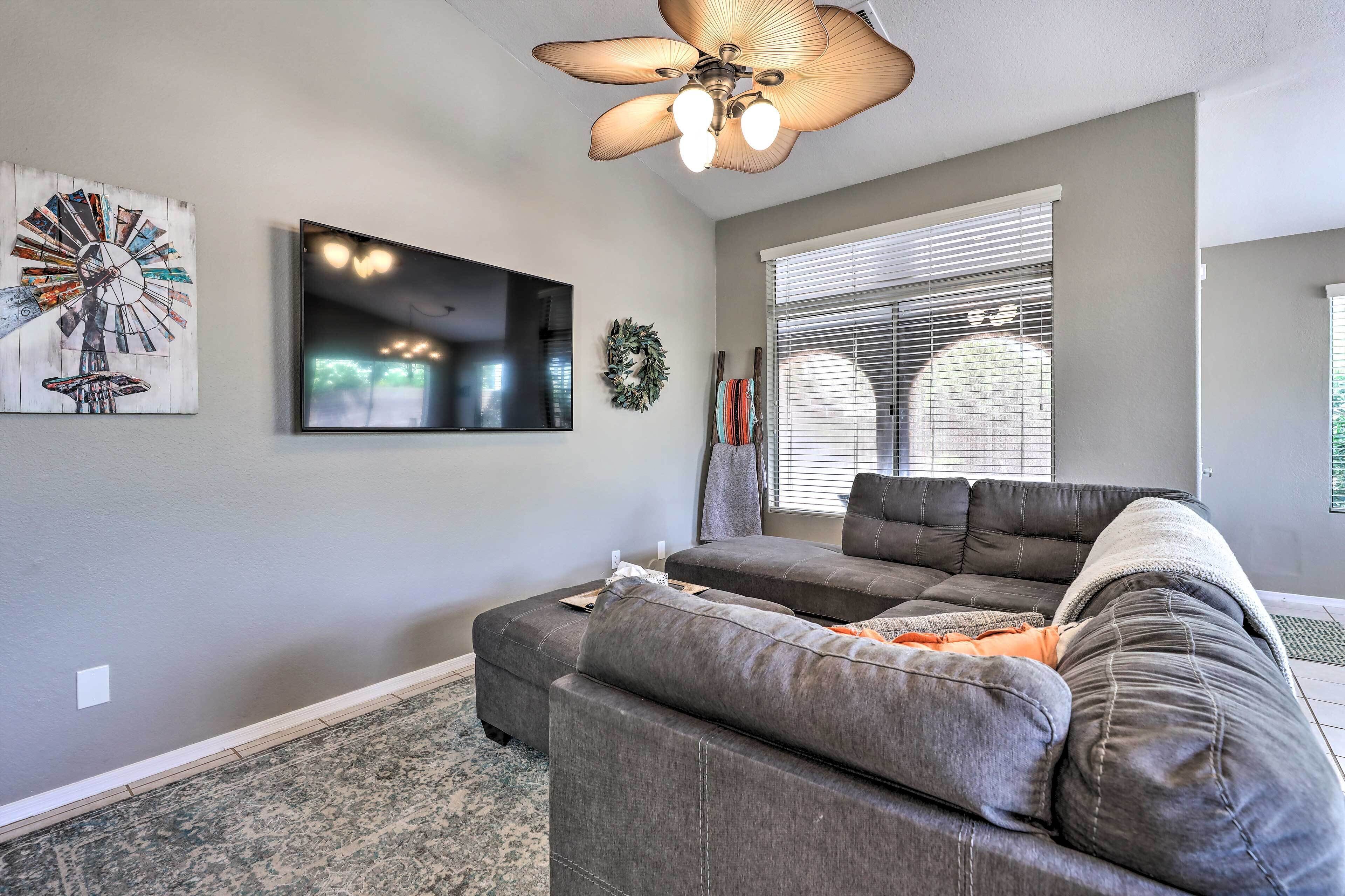 A living room with a gray sectional sofa, a ceiling fan, a flat-screen TV, and a large window with blinds.