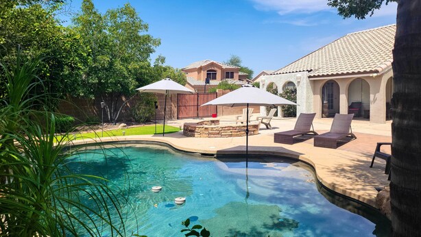 A swimming pool with lounge chairs and umbrellas, surrounded by a landscaped garden and a house with a tiled roof. - Beautiful Backyard: Comfortable Phoenix Getaway! (Phoenix)