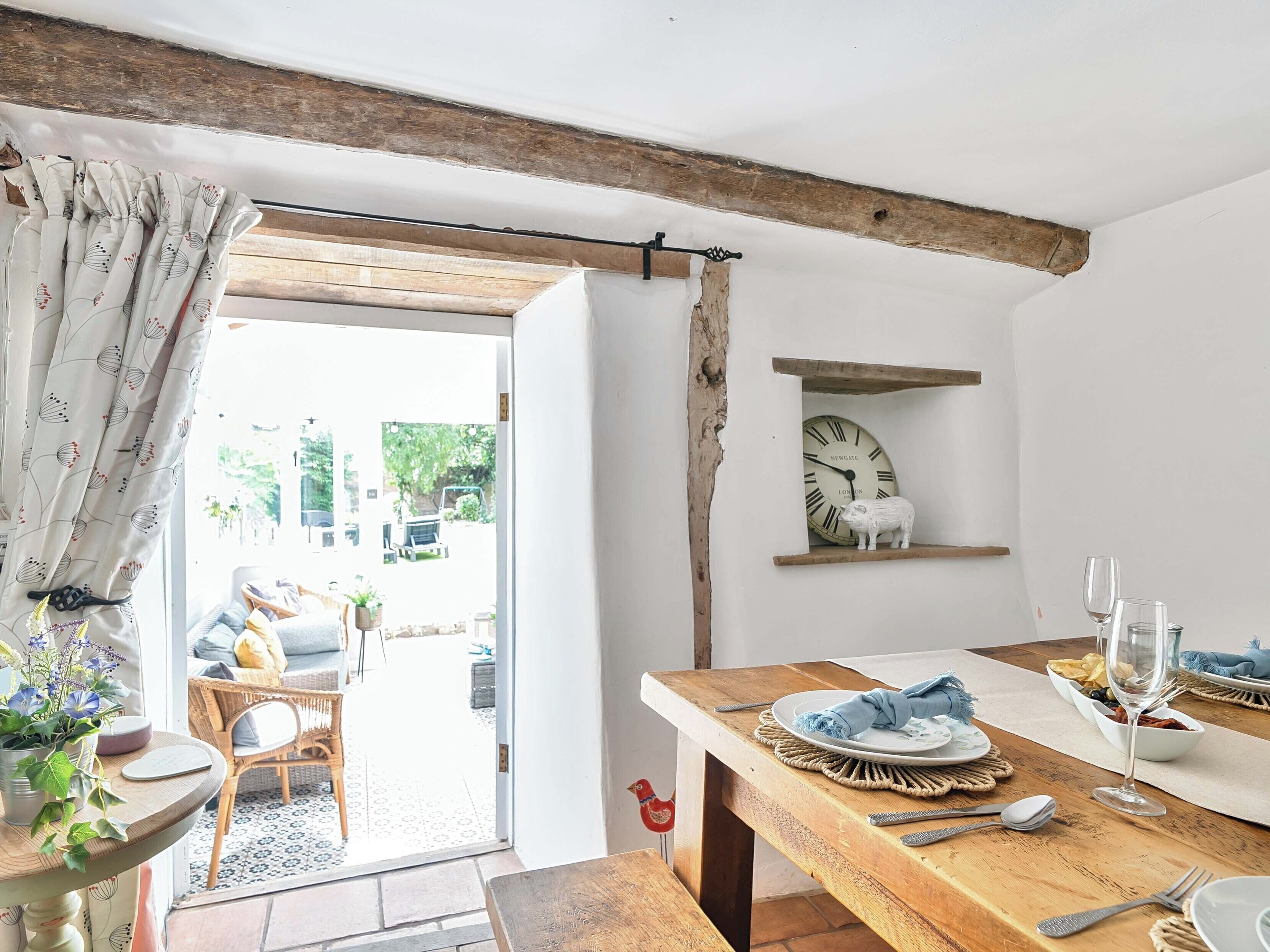 A dining area with a wooden table set for two, a wall clock, and a rustic shelf.