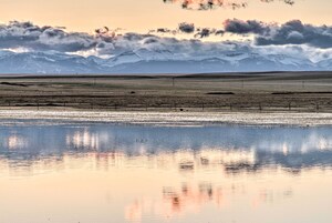 Uma paisagem extensa e aberta com um corpo d'água calmo refletindo o céu e montanhas ao longe.