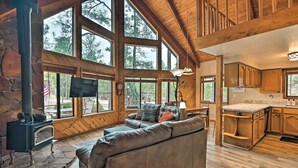 A spacious living area with a stone fireplace, large windows, and a kitchen with wooden cabinets. - 1 Mi to Lake: Secluded Cabin w/ Deck in Munds Park (Munds Park)