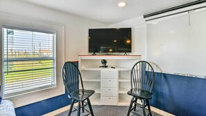 A room with a desk, two chairs, a flat-screen TV, a window with blinds, and a whiteboard. - Relaxing Surfside Bunkhouse - Walk to the Beach! (Freeport)